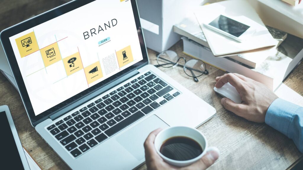 A laptop with the word Brand is displayed while someone works on the laptop and holds a mug of coffee, demonstrating brand identity.