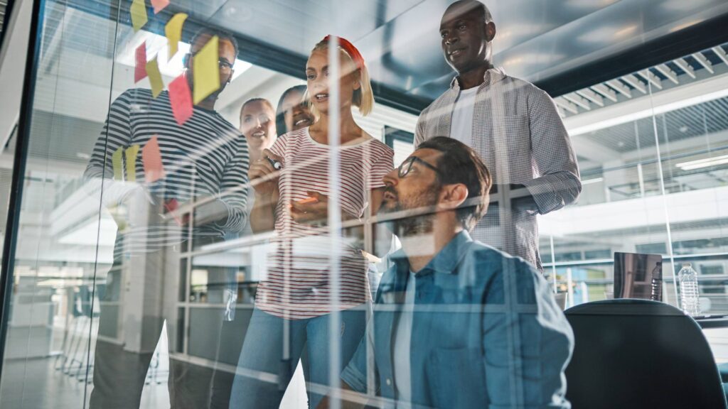 A group of team members brainstorm in front of a clear whiteboard with colorful post its, demonstrating entrepreneurship.