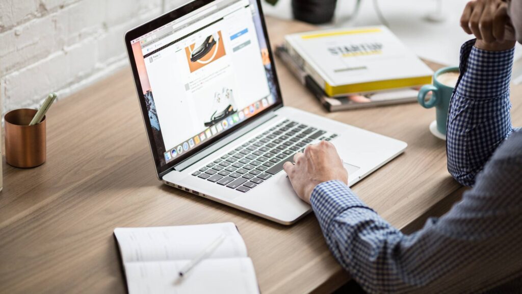 A person works on their laptop at a wooden desk with an open notebook to the left of his computer, demonstrating email marketing.