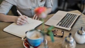 A person sits at a desk while writing in a notebook with a laptop on the side, demonstrating SEO keyword research for 2025.